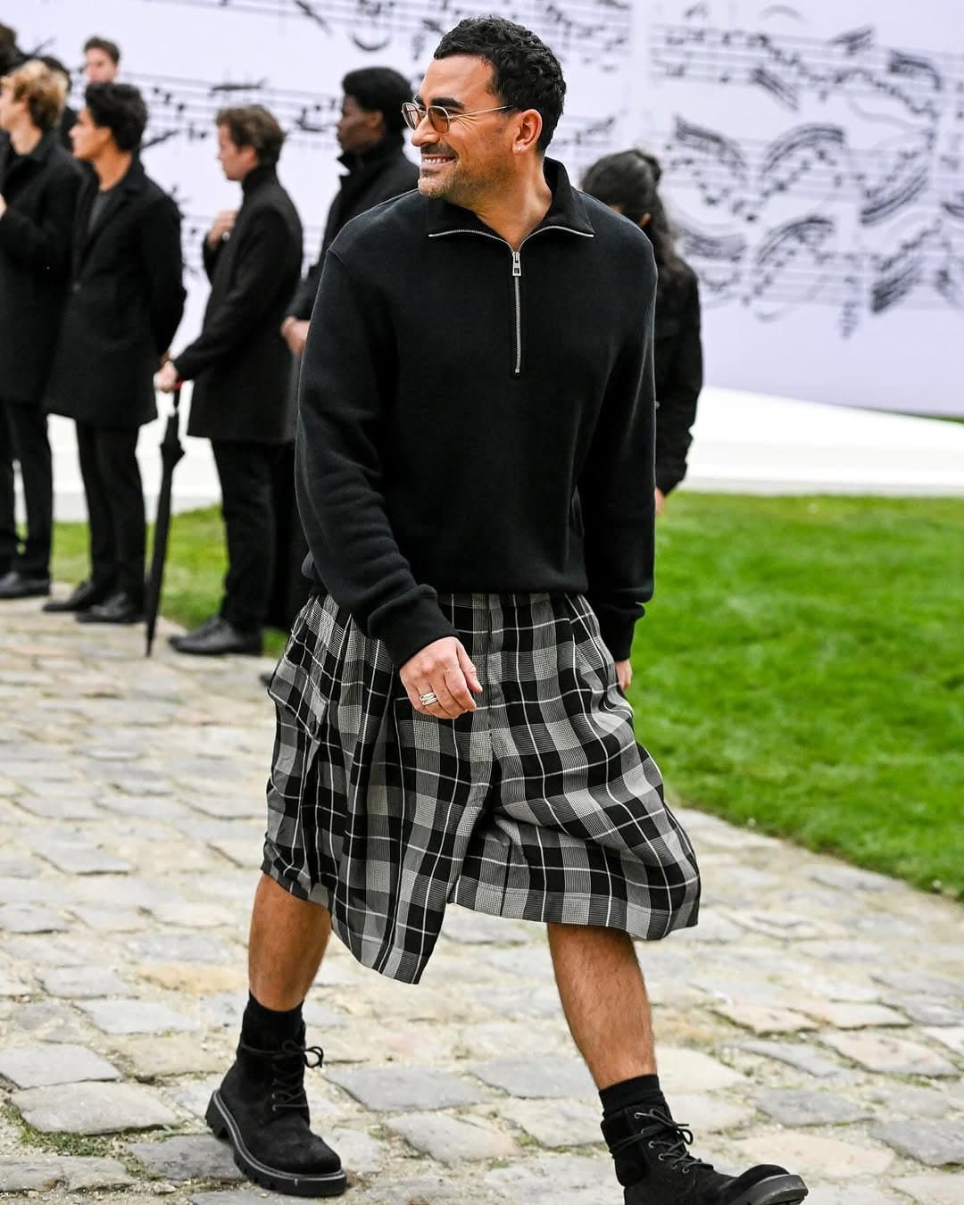 A black half-zip pullover is styled with voluminous black-and-grey plaid knee-length shorts, black socks, and black lace-up boots.