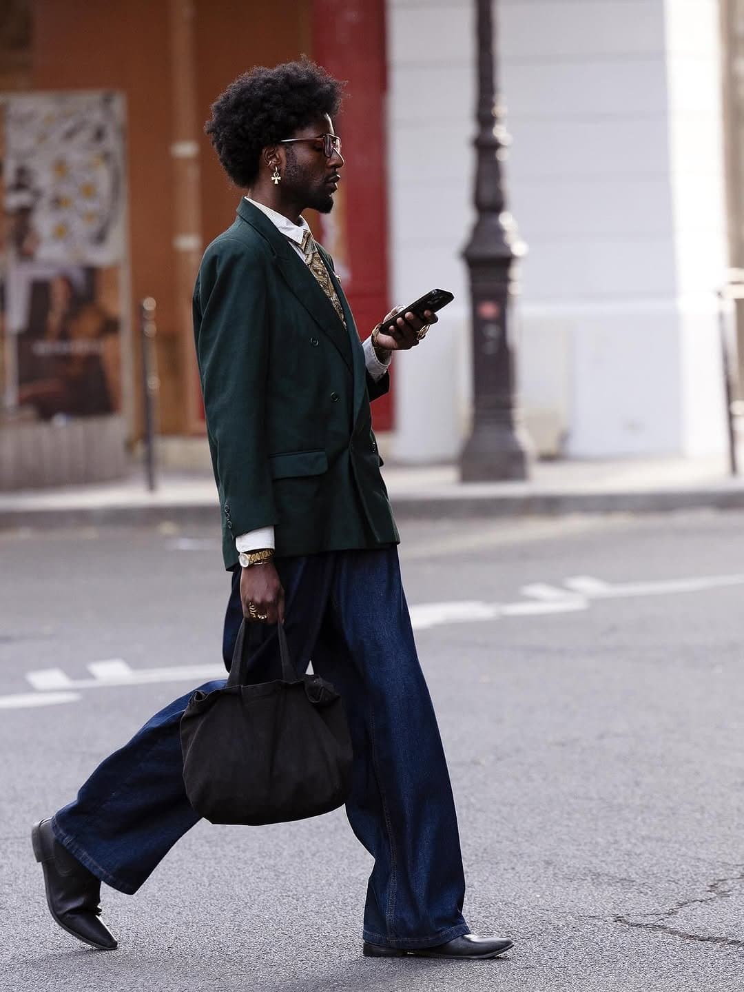 A dark green double-breasted blazer is styled with a striped shirt, patterned tie, wide dark denim trousers, black boots, and a black tote.