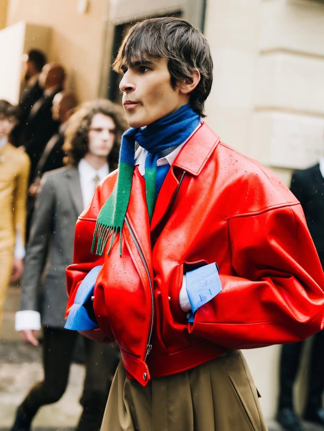 A bright red oversized leather jacket is layered over blue shirting, a blue-green scarf, and pleated tan trousers.