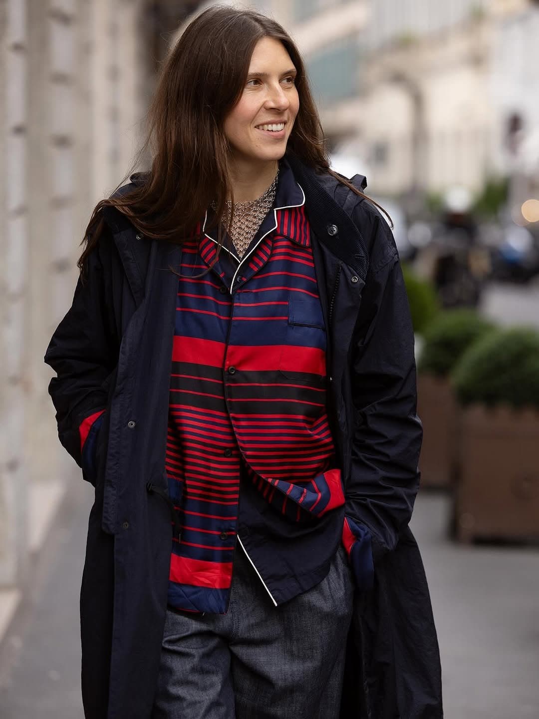 A navy outer coat layers over a red-and-navy striped shirt with grey trousers and a chunky silver chain necklace.