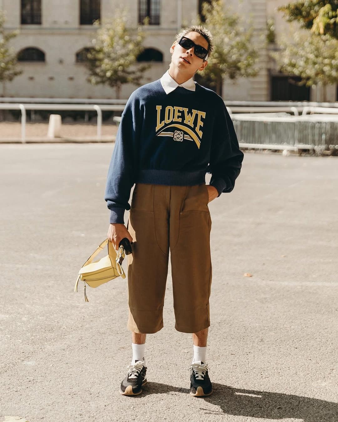 A navy logo sweatshirt over a white collared shirt is styled with brown cropped utility shorts, white socks, black sneakers, sunglasses, and a pale yellow handbag.
