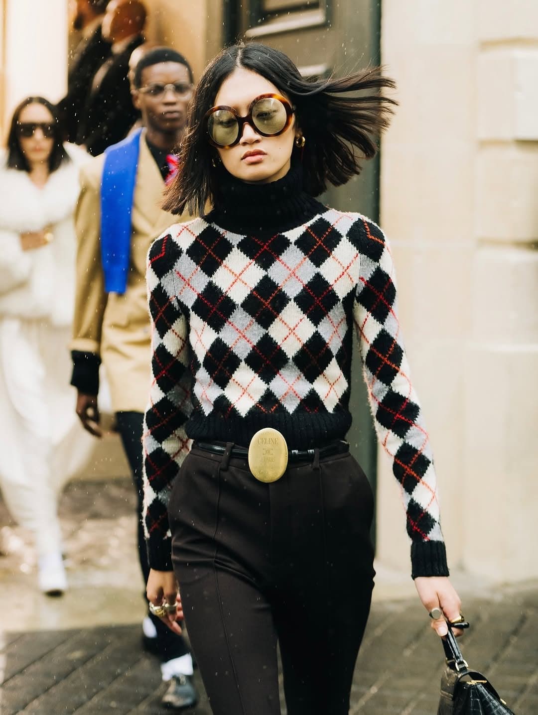 A black turtleneck layered under a cropped argyle sweater with high-waisted black trousers, oversized round sunglasses, and a statement oval belt buckle.