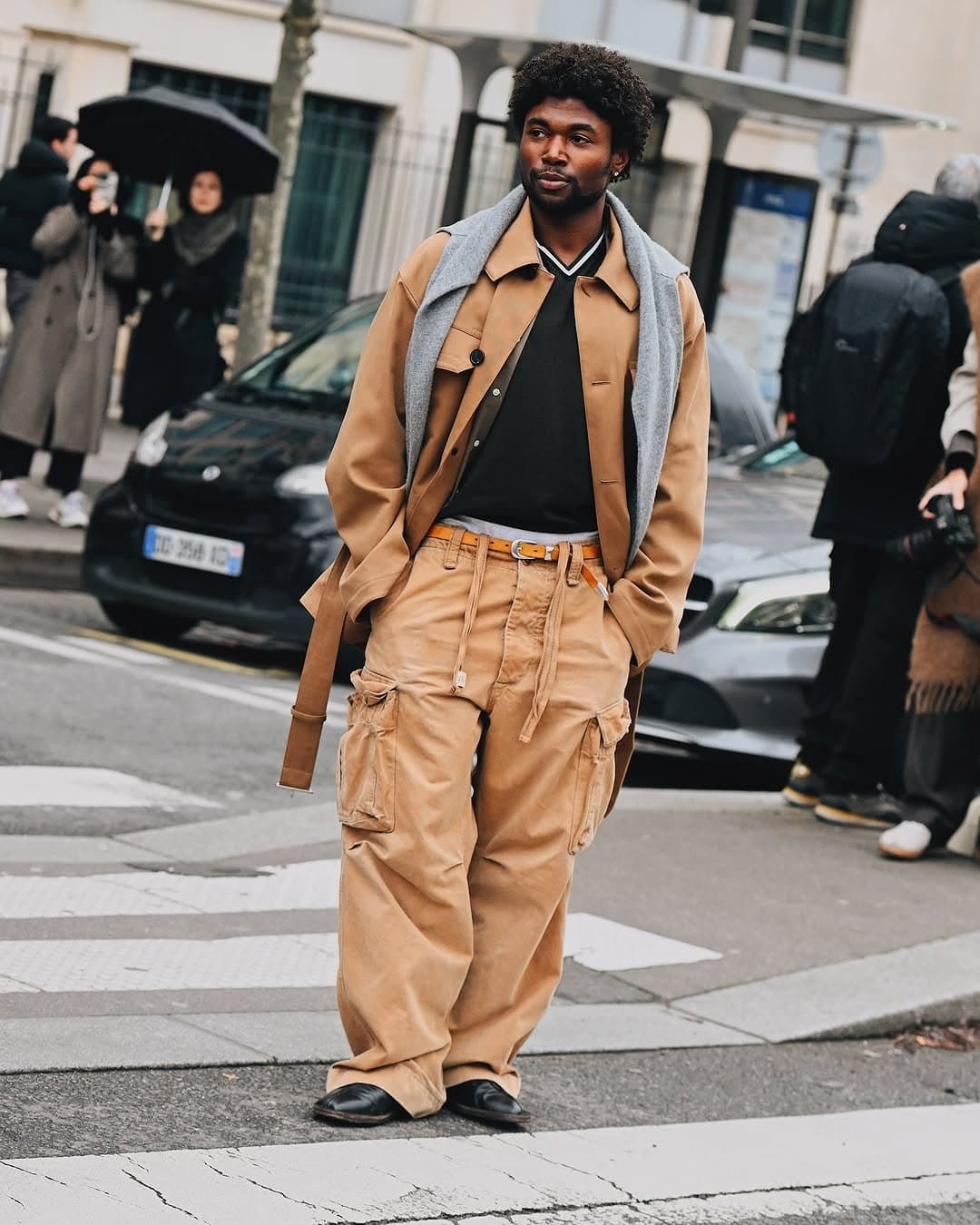 A tan utility jacket and oversized tan cargo trousers are styled with a black knit top, grey sweater draped over the shoulders, tan belt, and black leather shoes.
