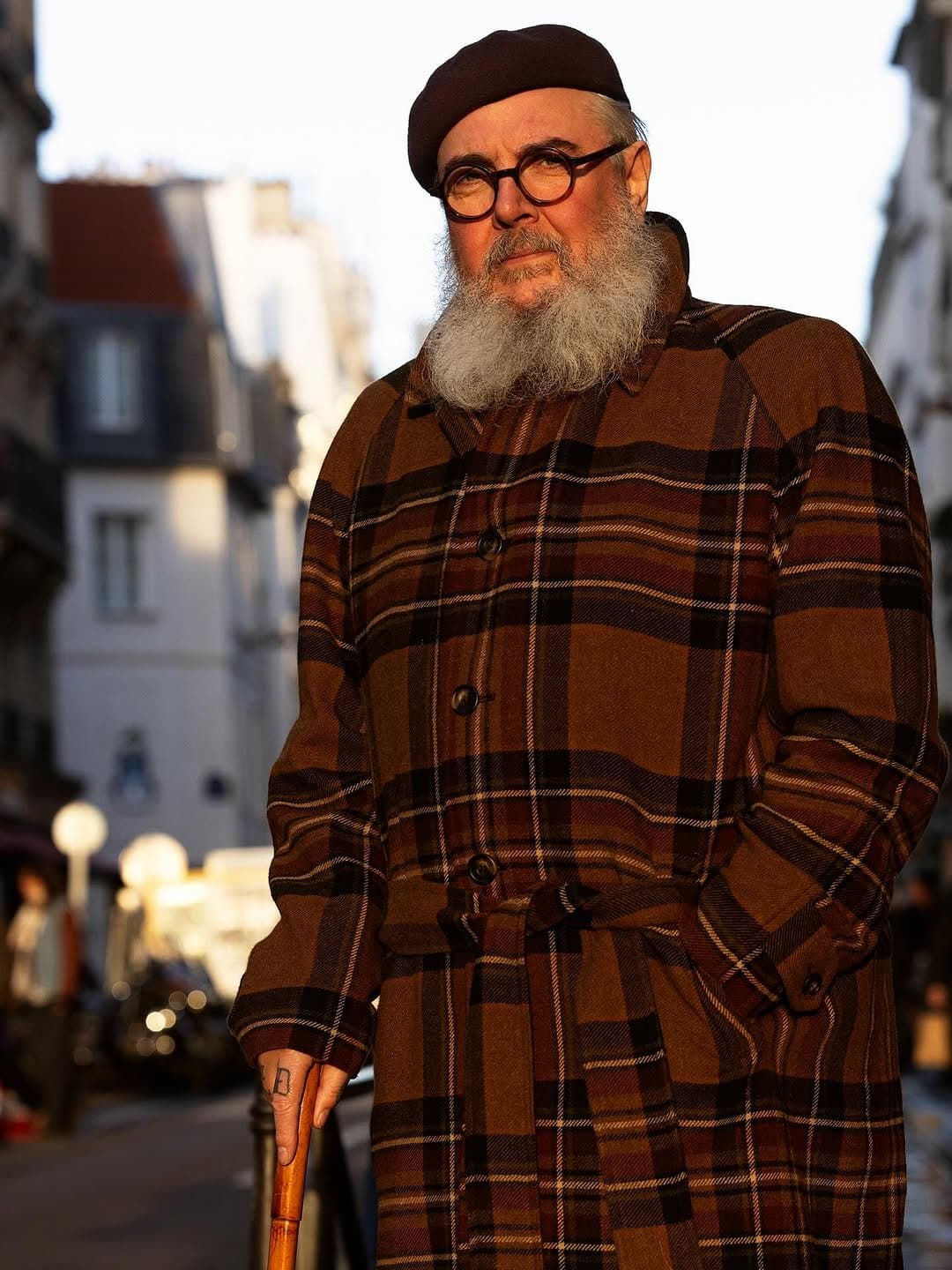 A brown plaid belted overcoat is styled with a dark beret and round glasses.