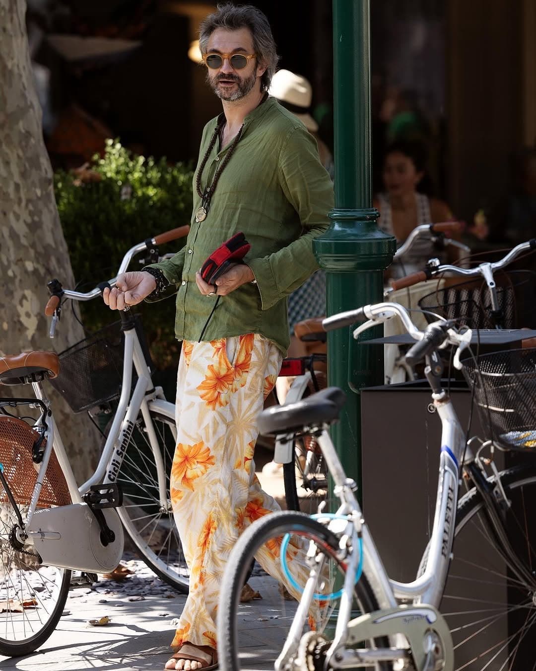 A relaxed olive shirt is styled with cream floral trousers, sandals, tinted round sunglasses, and layered necklaces.