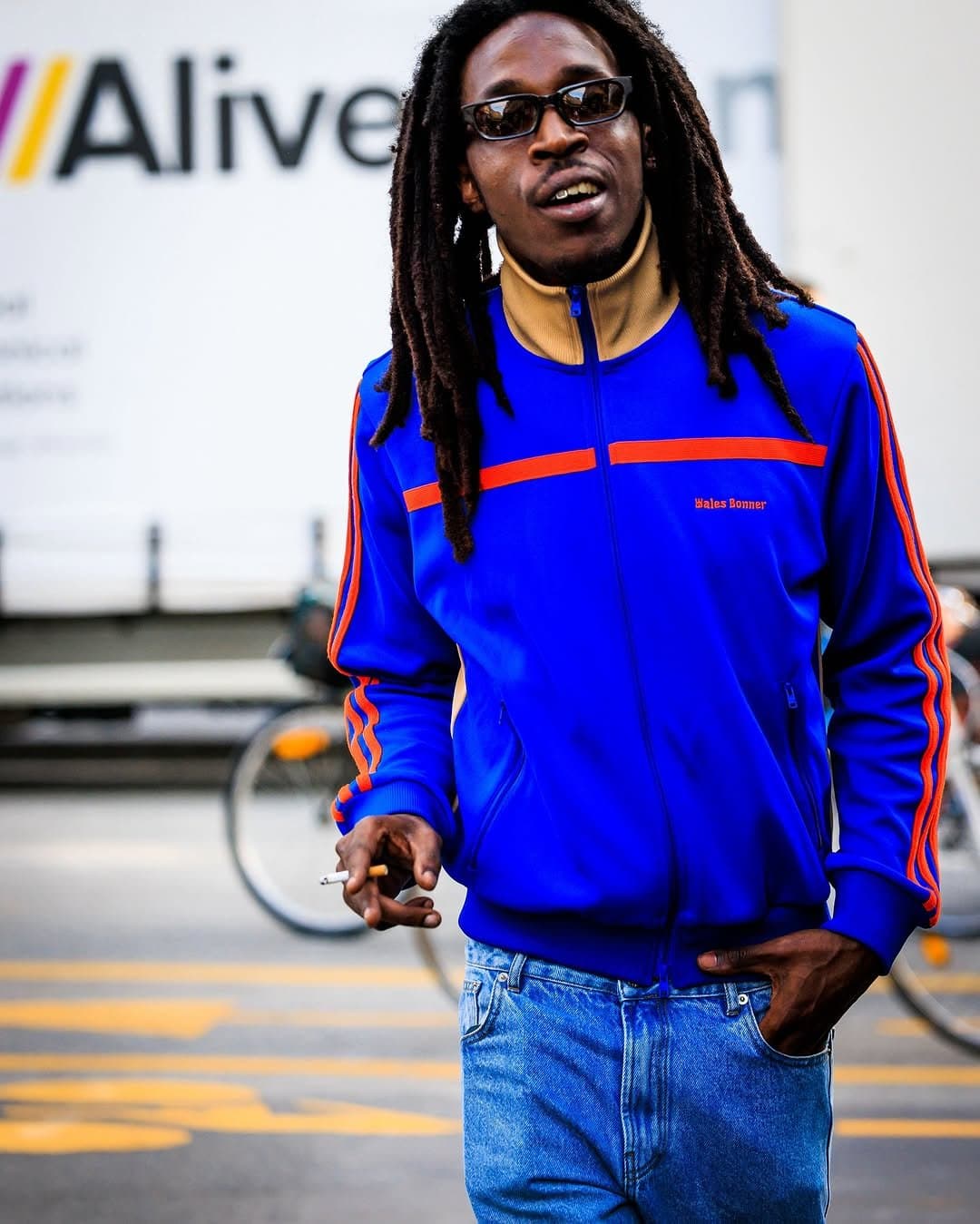 A bright blue track jacket with orange striping is worn over light blue jeans and narrow black sunglasses.