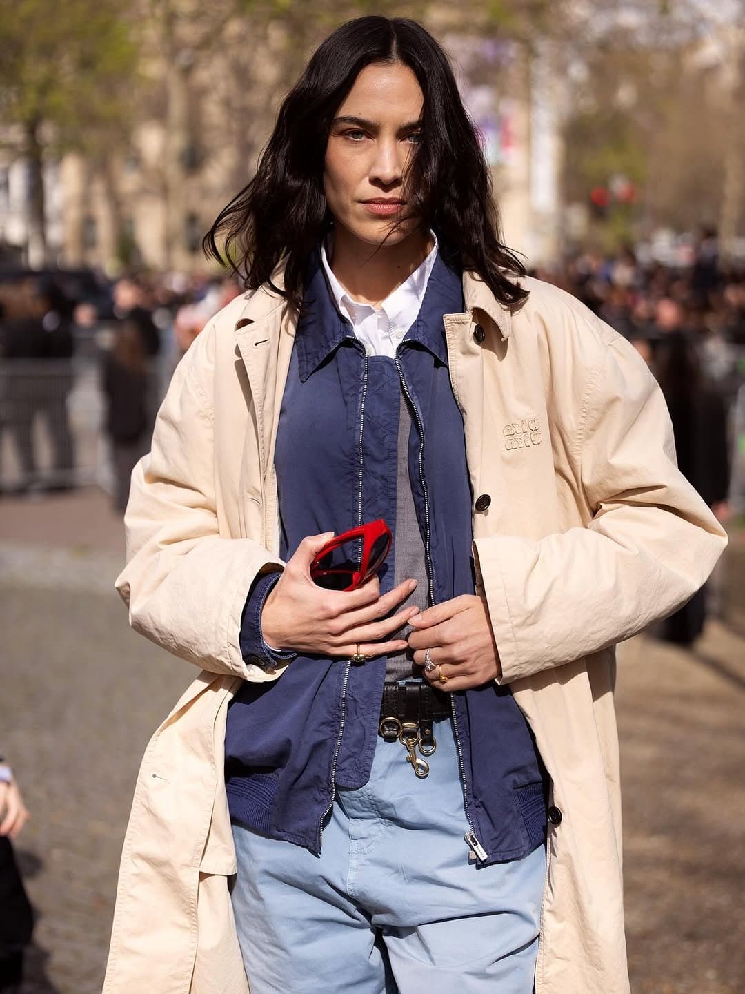 A cream oversized coat is layered over a navy zip jacket, white shirt, grey top, and pale blue trousers with a black belt and rings.