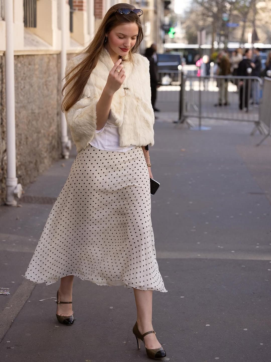 Cream faux-fur cropped jacket, white top, polka-dot midi skirt, and two-tone Mary Jane heels on a city sidewalk.