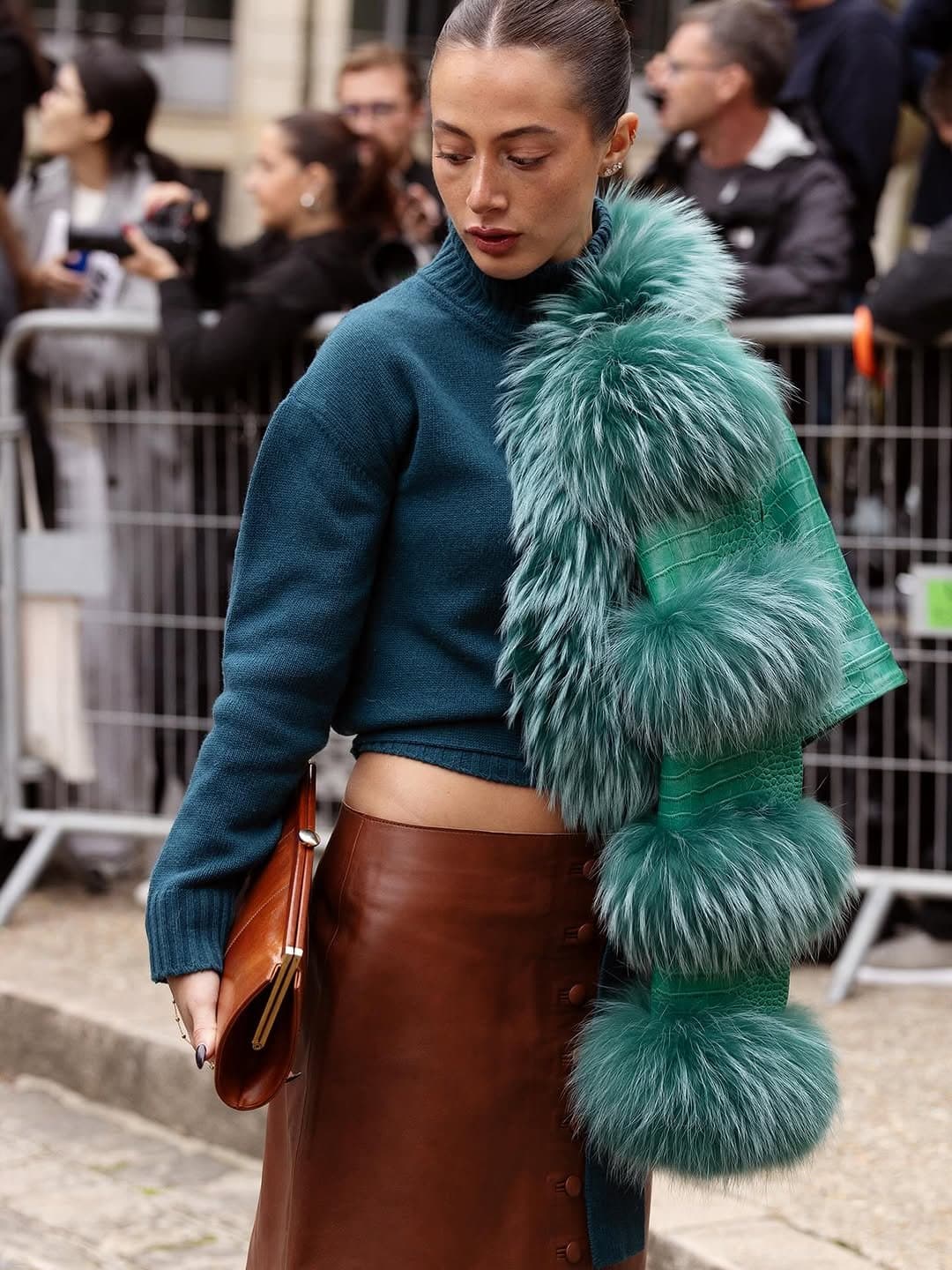A teal cropped knit sweater is styled with a brown leather skirt, a fluffy teal fur-trimmed scarf, and a cognac clutch.