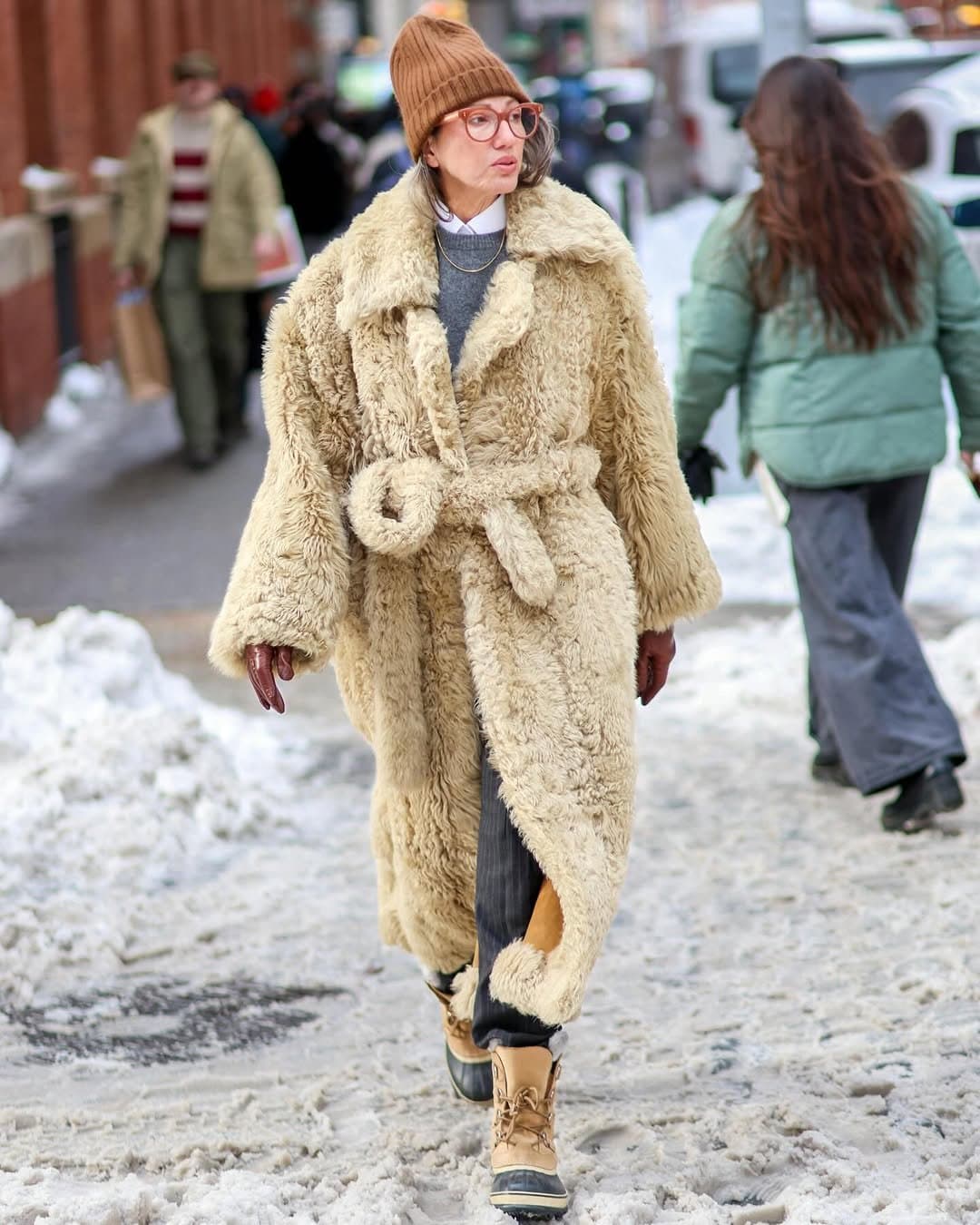 A plush cream belted shearling coat is styled with a brown ribbed beanie, layered shirting and knitwear, dark trousers, brown gloves, and tan winter boots.