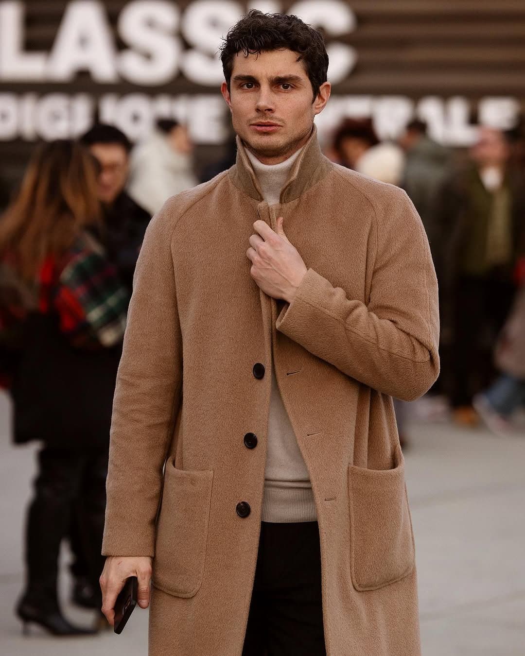 A camel textured overcoat is layered over a light ribbed turtleneck and dark trousers.