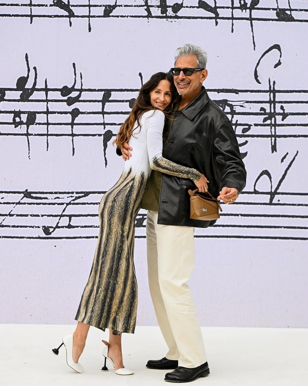 A woman in a white-and-black patterned midi dress and white heels embraces a man in a black leather jacket, olive top, cream trousers, black loafers, and sunglasses.