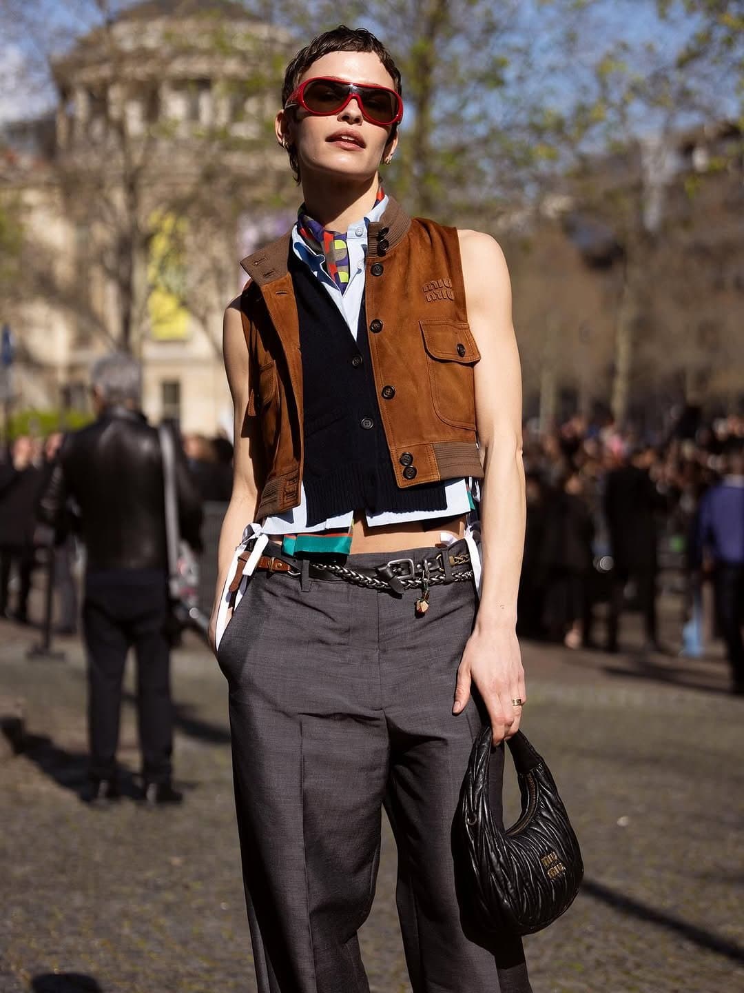 A cropped layered vest-and-shirt look with grey wide trousers, red sunglasses, chain belt, and a black quilted handbag.