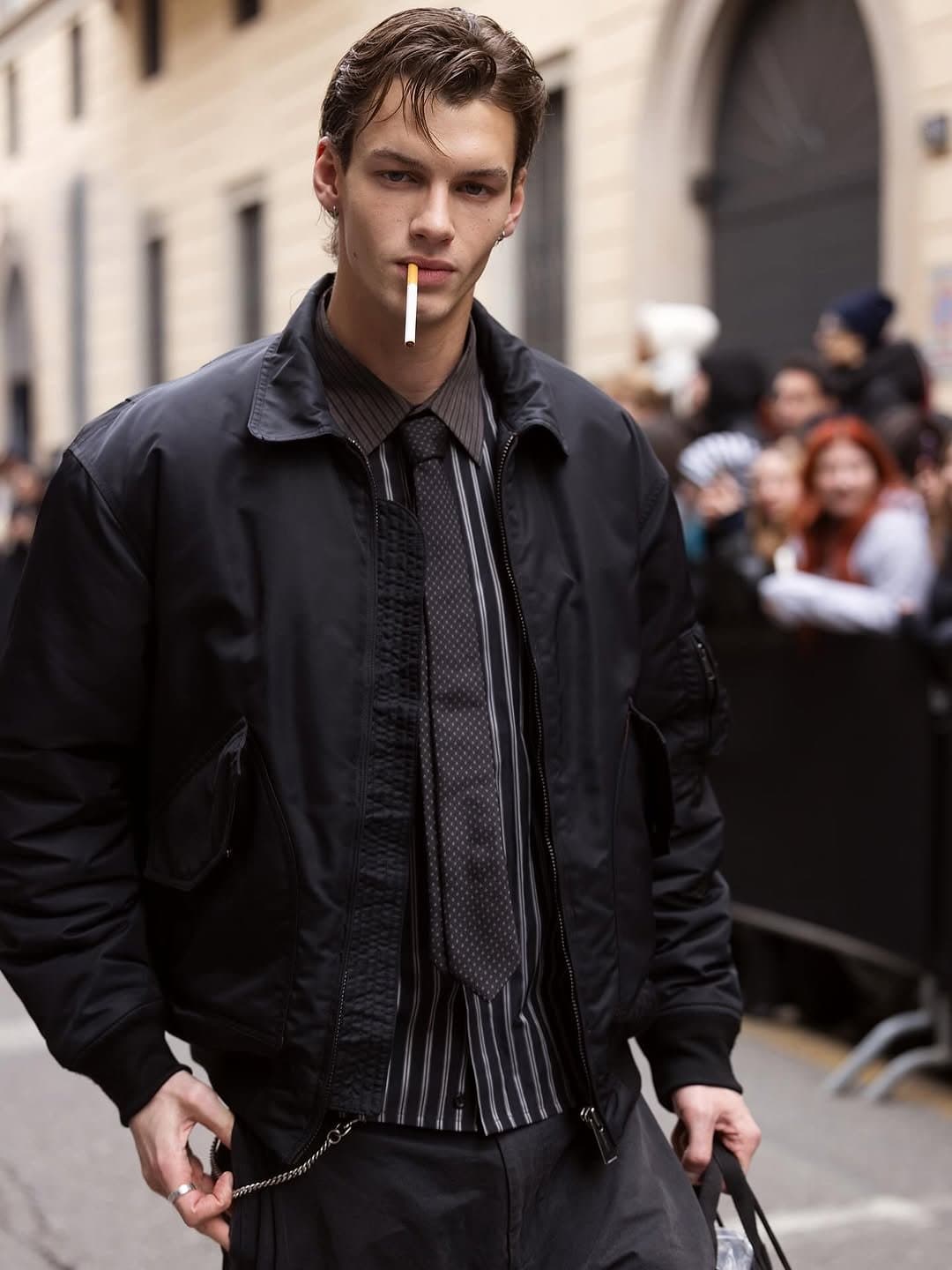A black cropped bomber is layered over a striped shirt, dark tie, textured black knit, and dark trousers with a chain accent.