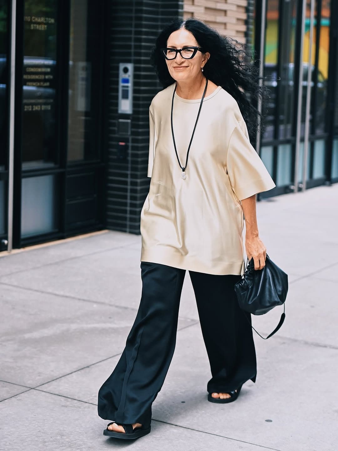 An oversized cream tunic top is styled with wide black trousers, black sandals, a black pouch bag, and a long pendant necklace.
