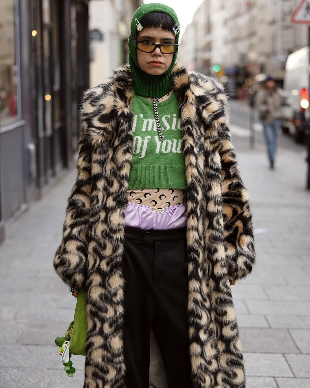 A green hooded knit crop top is layered under a long animal-print faux-fur coat with black trousers and visible lavender underwear styling.
