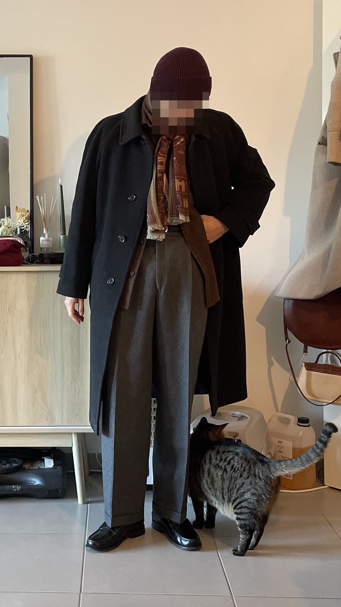 A long black overcoat is layered over tailored grey wide-leg trousers, a patterned burgundy scarf, and glossy black loafers.