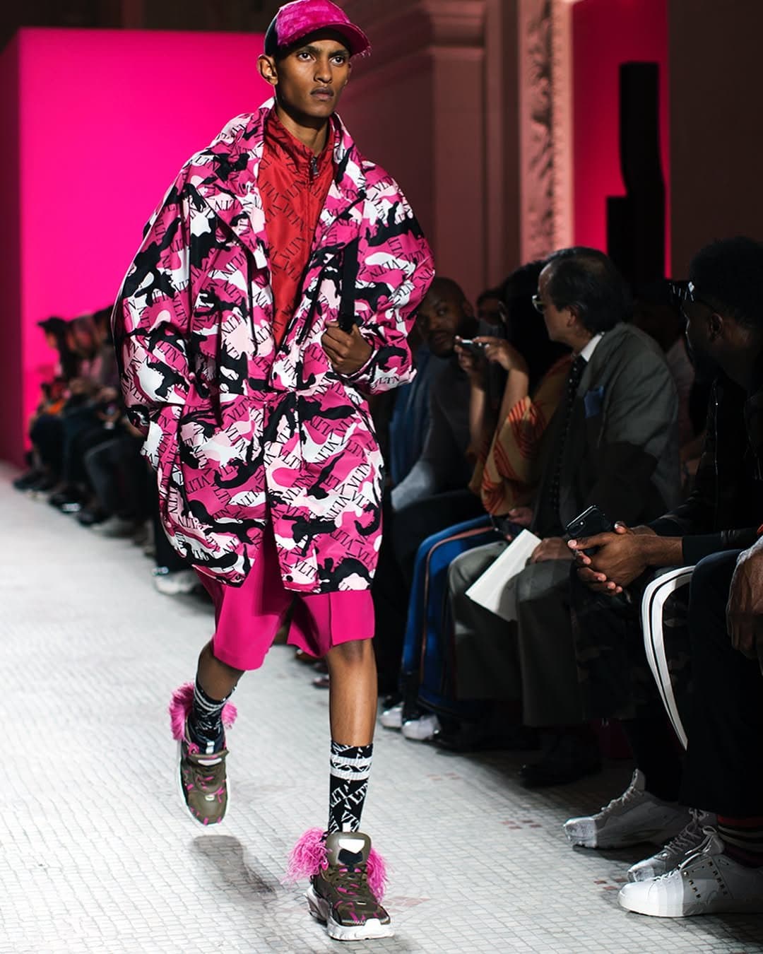 A runway look pairs a pink logo-camouflage oversized coat with magenta shorts, printed socks, chunky sneakers, and a matching cap.
