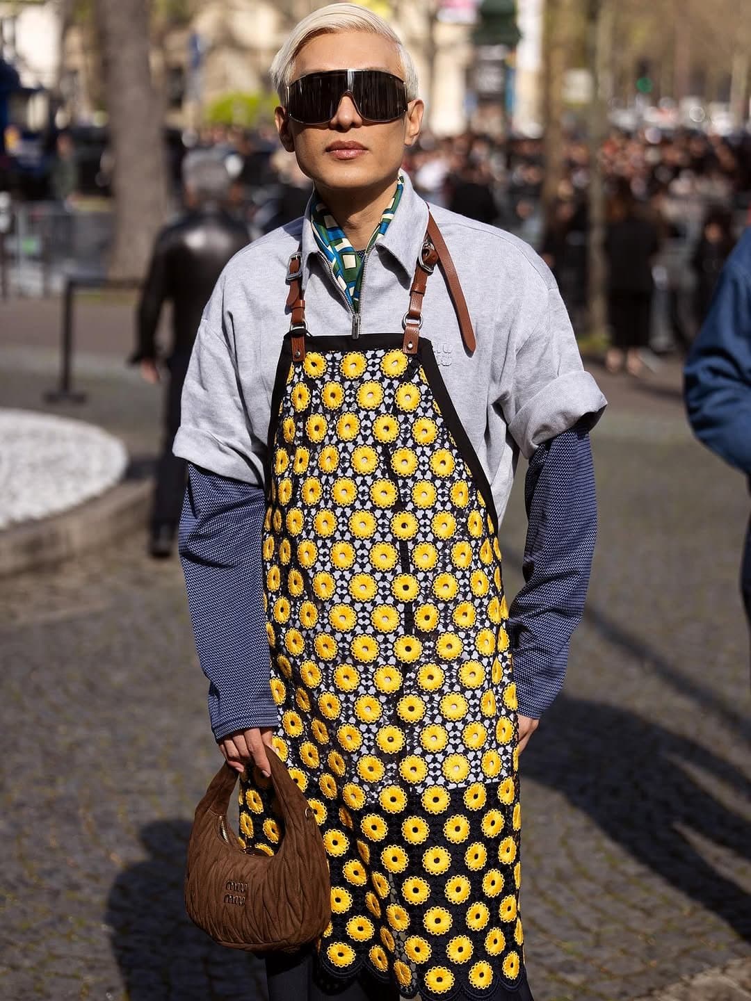 A sunflower-patterned apron dress is layered over a pale grey short-sleeve zip shirt, blue long sleeves, oversized visor sunglasses, and a brown quilted handbag.