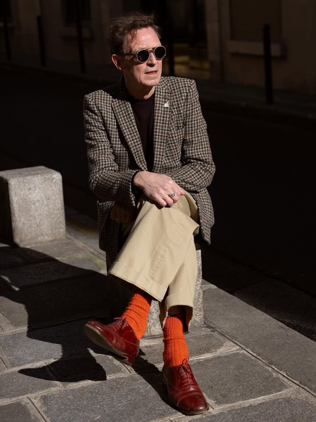 A houndstooth blazer is styled with a dark knit top, beige cropped trousers, bright orange socks, and burgundy leather lace-up shoes.