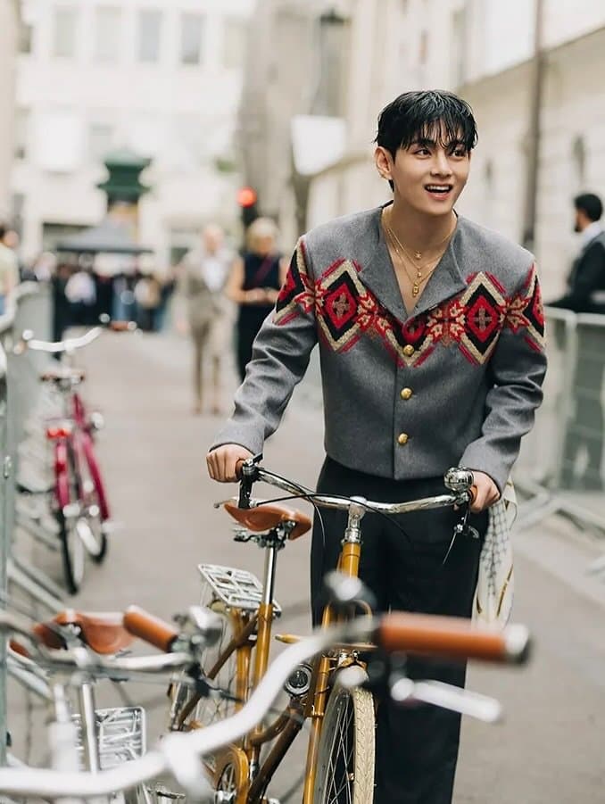 A grey cropped cardigan with red geometric embroidery is styled open at the neck with black trousers and layered gold jewelry.
