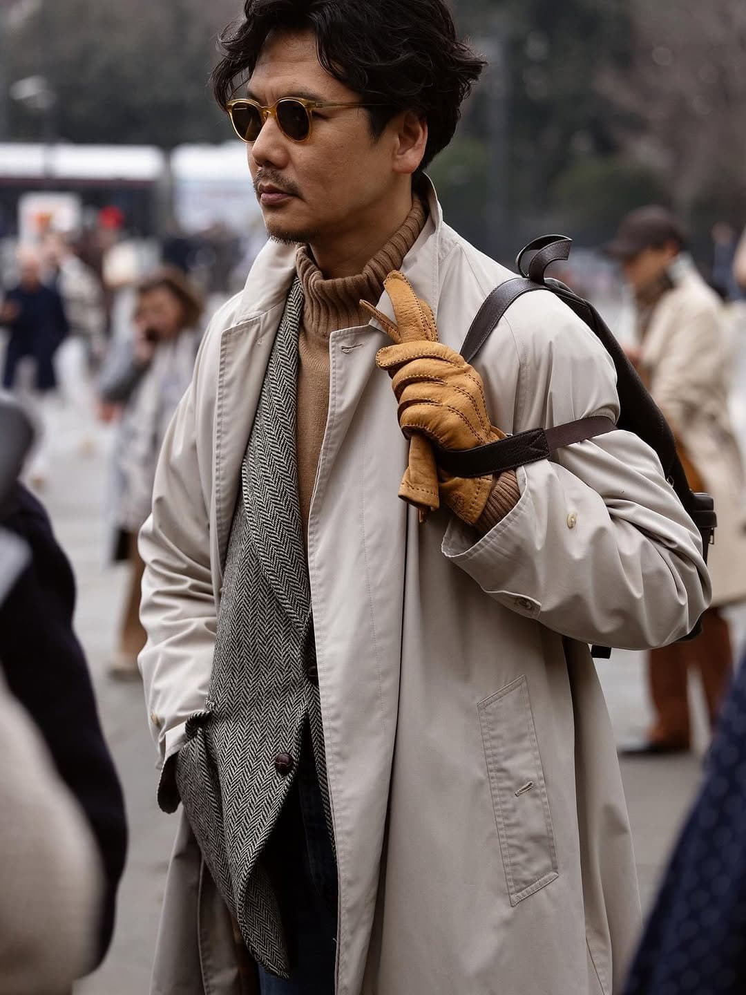 A layered neutral street-style look with a light trench coat, camel turtleneck, herringbone tailoring, leather gloves, sunglasses, and a dark shoulder bag.
