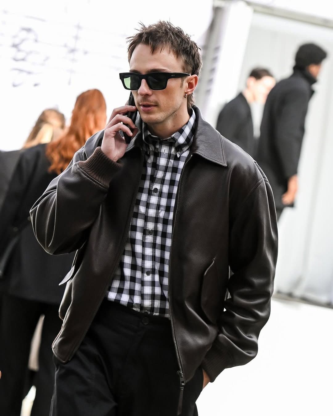 A dark leather blouson jacket is worn over a black-and-white checked shirt with black trousers and rectangular sunglasses.